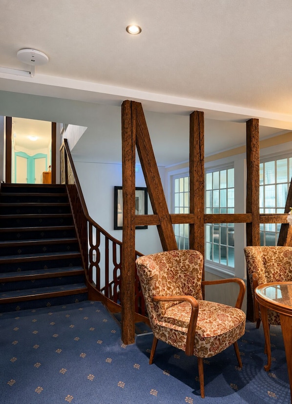Una habitación con alfombra estampada, sillas de madera, una escalera con barandilla y una ventana con persianas.