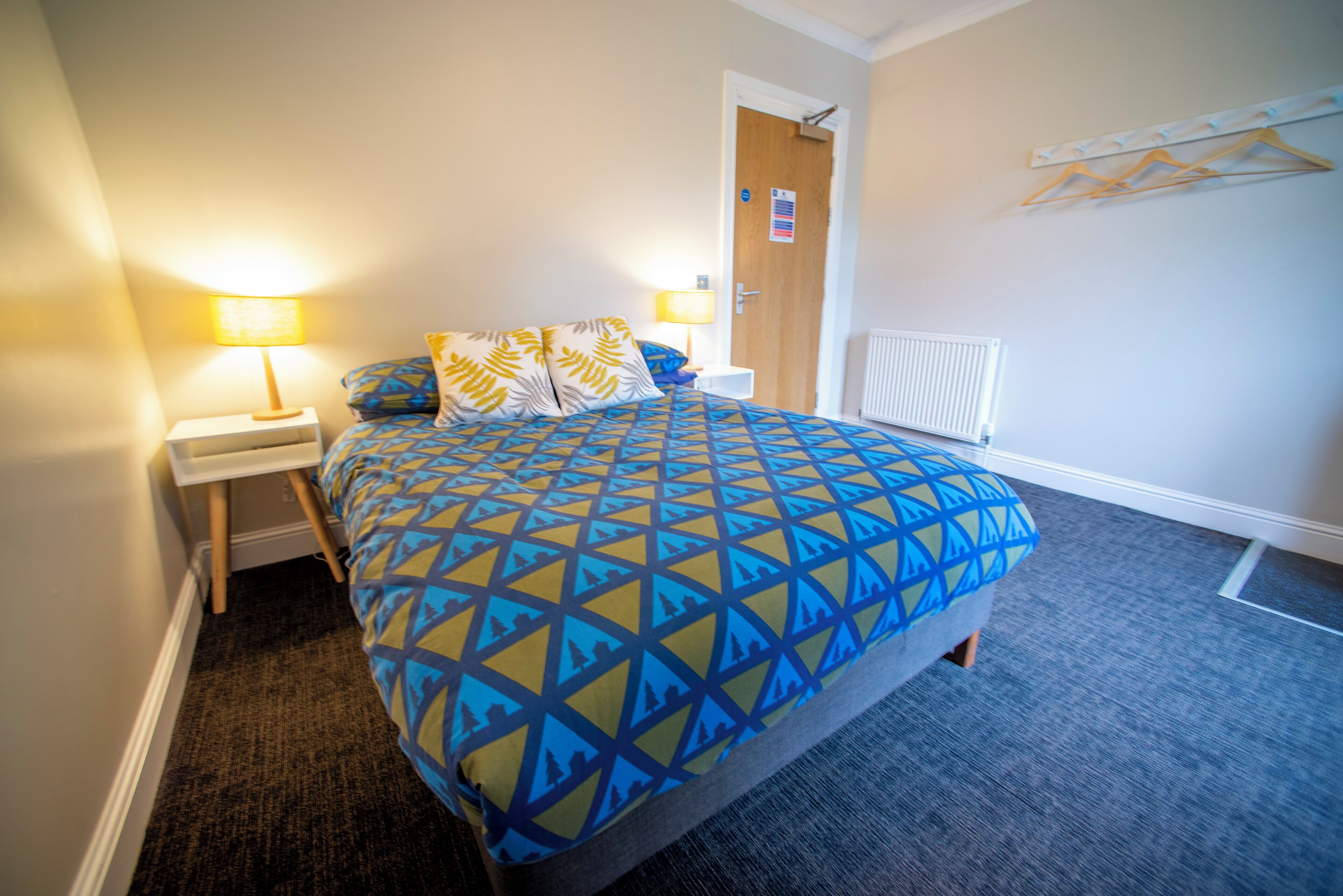 A hotel room with a large bed, two bedside lamps, a radiator, and a coat rack.