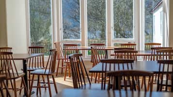 Un comedor con varias mesas y sillas de madera, ventanas amplias con vistas a árboles, y una puerta a la derecha.