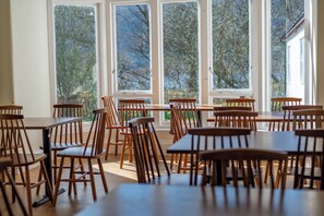 A dining area with multiple wooden tables and chairs, large windows with a view of trees, and a door to the right. - Rowardennan Youth Hostel (Balmaha)