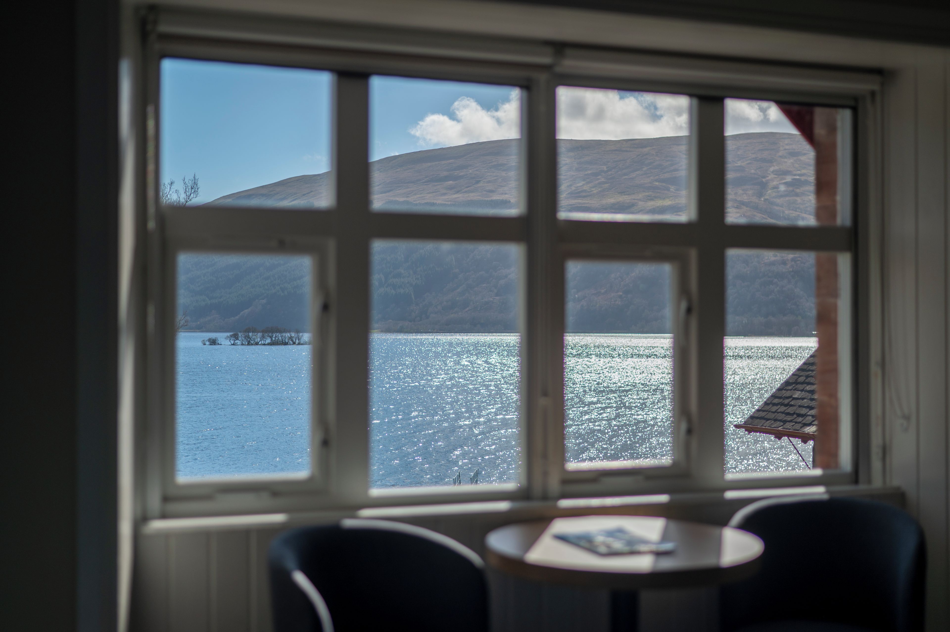 Habitación con un gran ventanal con vistas a un lago y montañas. En primer plano, una pequeña mesa con sillas.