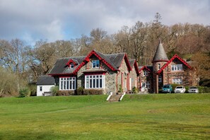 A stone building with red accents and a conical roof, surrounded by a grassy field and trees. - Rowardennan Youth Hostel (Balmaha)