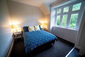 A bedroom with a bed, two bedside lamps, and a window with a view of greenery. - Rowardennan Youth Hostel (Balmaha)