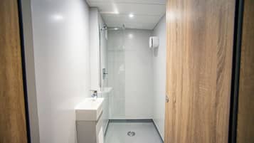 A narrow, modern bathroom with a glass shower enclosure, a white sink, and a wooden door.