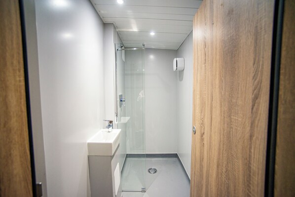 Un baño moderno y estrecho con cabina de ducha de vidrio, un lavabo blanco y una puerta de madera.