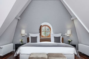 Un dormitorio con techo inclinado, una cama con ropa de cama blanca, dos mesitas de noche con lámparas y una ventana con marco de madera.
