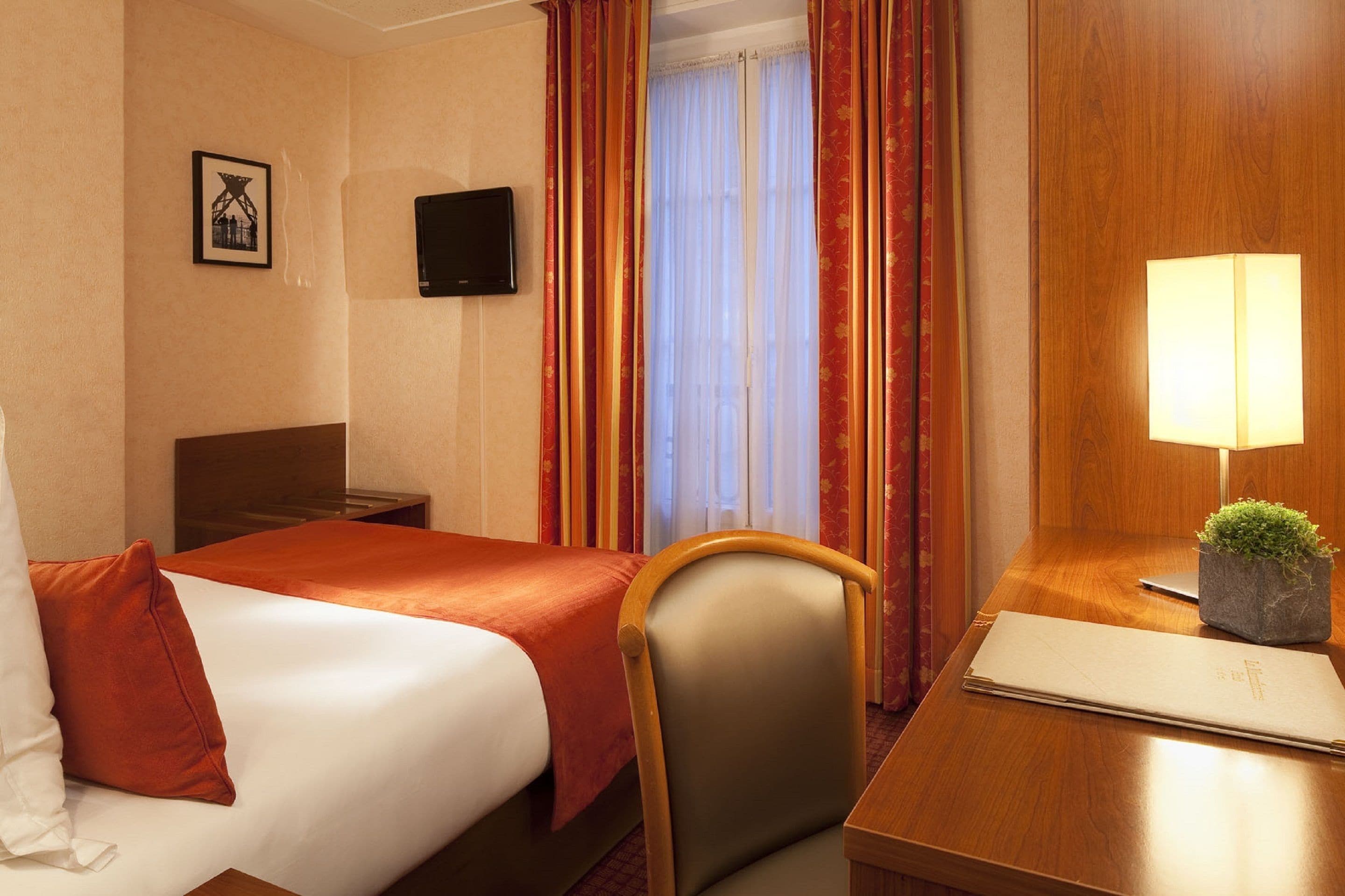 A hotel room with a bed, a desk with a lamp, a chair, a TV, and a window with curtains.