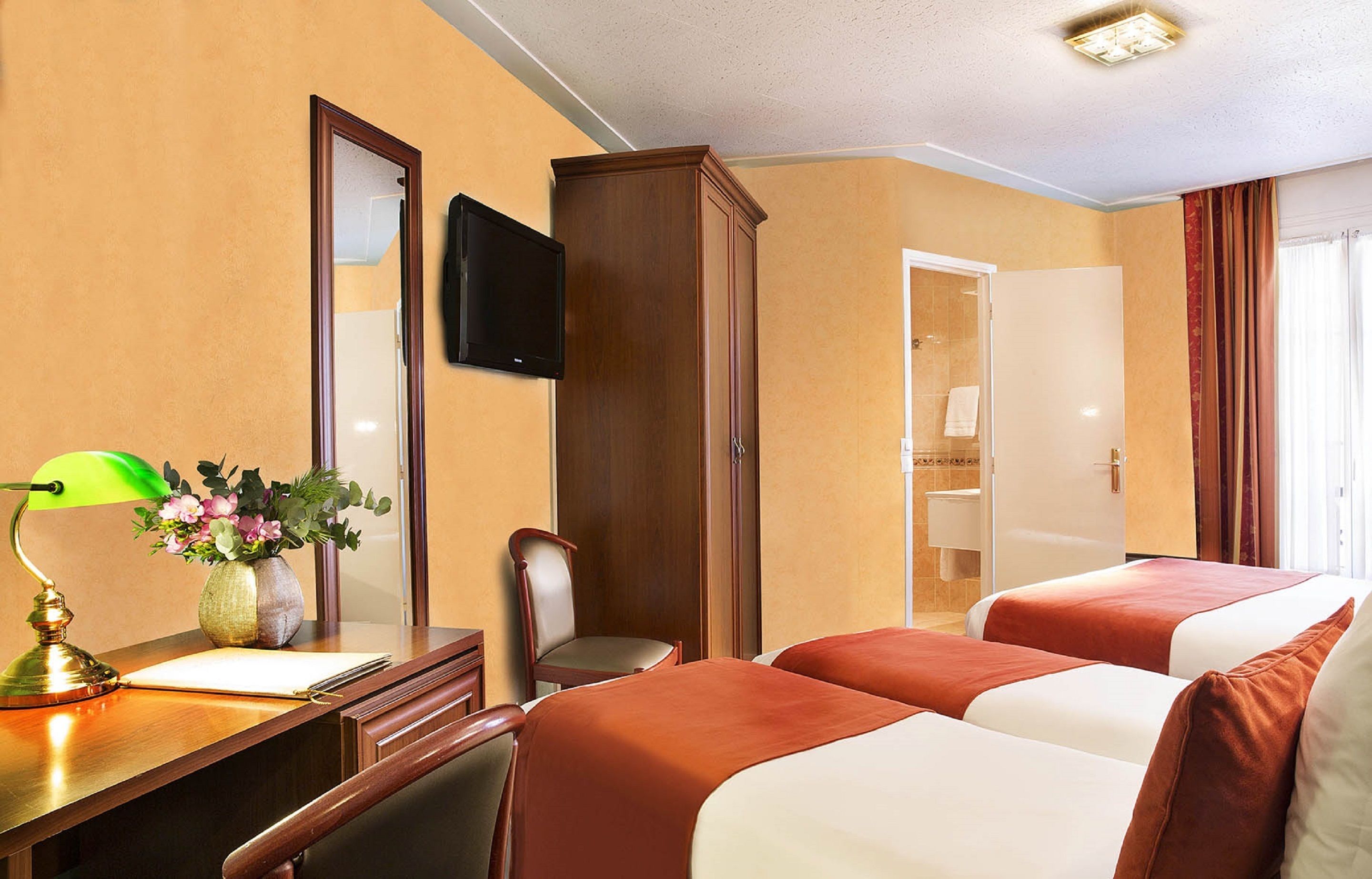 A hotel room with two beds, a desk with a lamp, a chair, a TV, and a bathroom door.