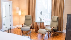 Une chambre d’hôtel avec un lit, deux fauteuils, une petite table ronde, une lampe et une vue sur l’extérieur grâce à de grandes fenêtres.