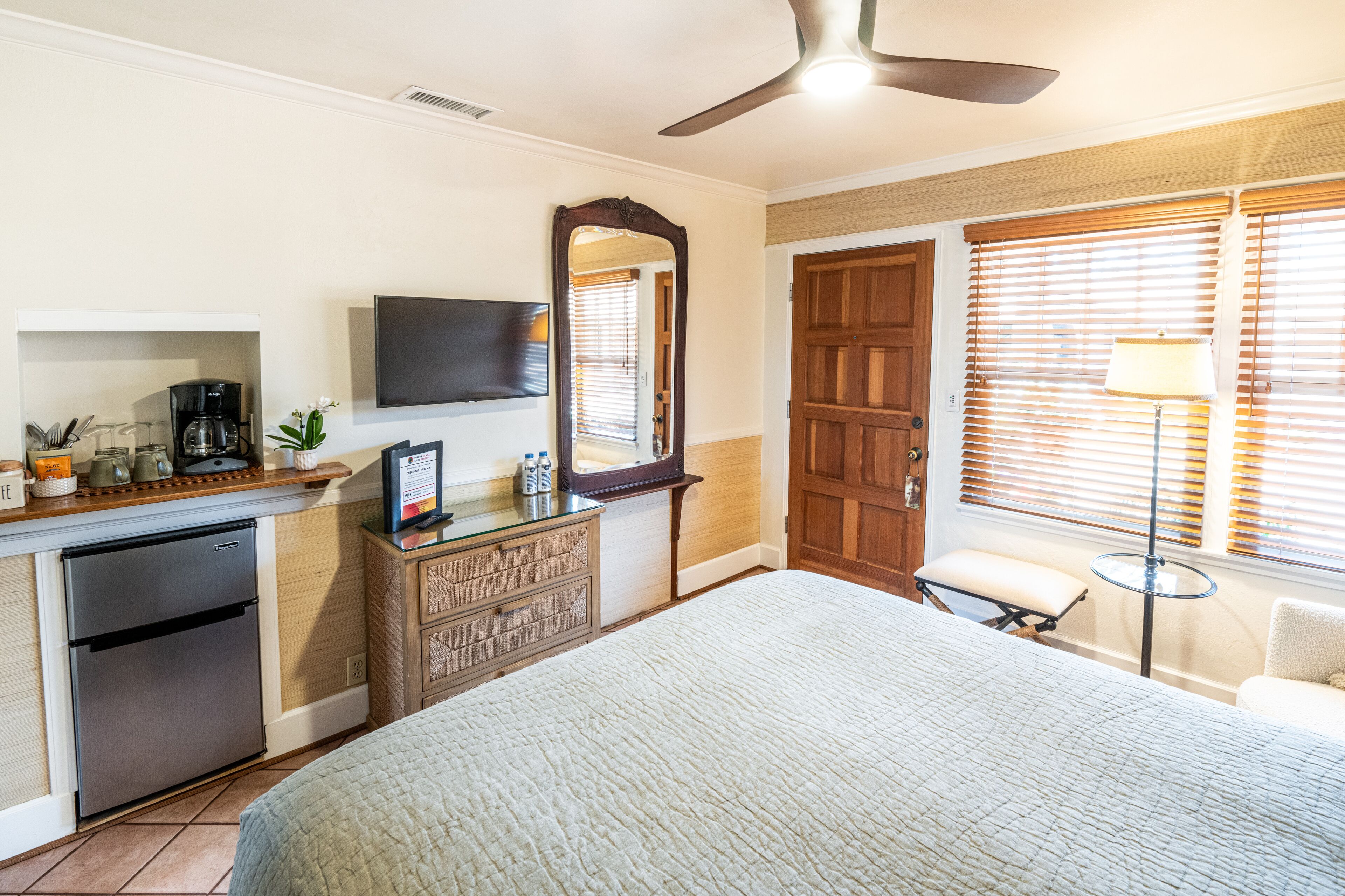 A hotel room with a bed, a desk with a refrigerator, a mirror, a television, a window with blinds, and a ceiling fan.