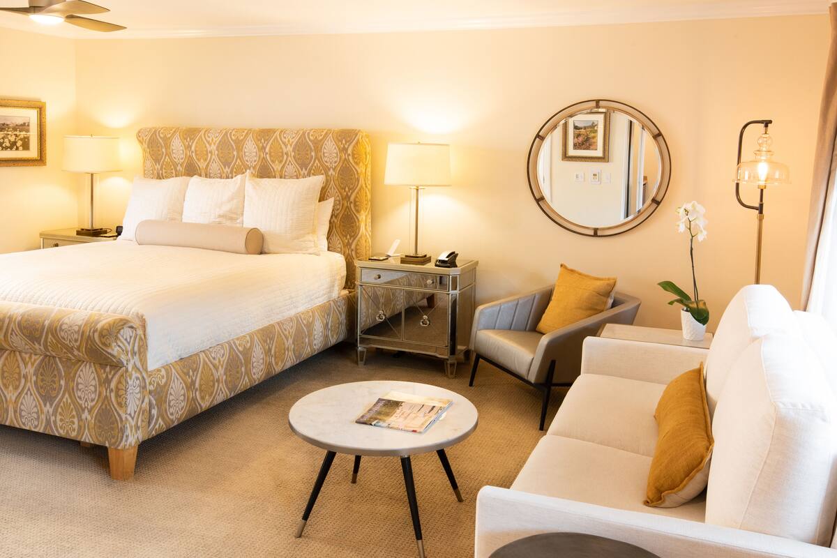 Ein Hotelzimmer mit einem Bett, zwei Nachttischen mit Lampen, einem runden Spiegel, einem kleinen runden Tisch mit einer Zeitschrift, einem Sofa und einer Topfpflanze.