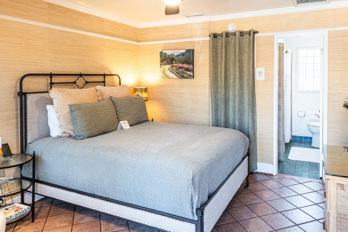 Ein ordentlich bezogenes Bett mit grauer Decke, ein Nachttisch mit Lampe und ein Gemälde an der Wand.