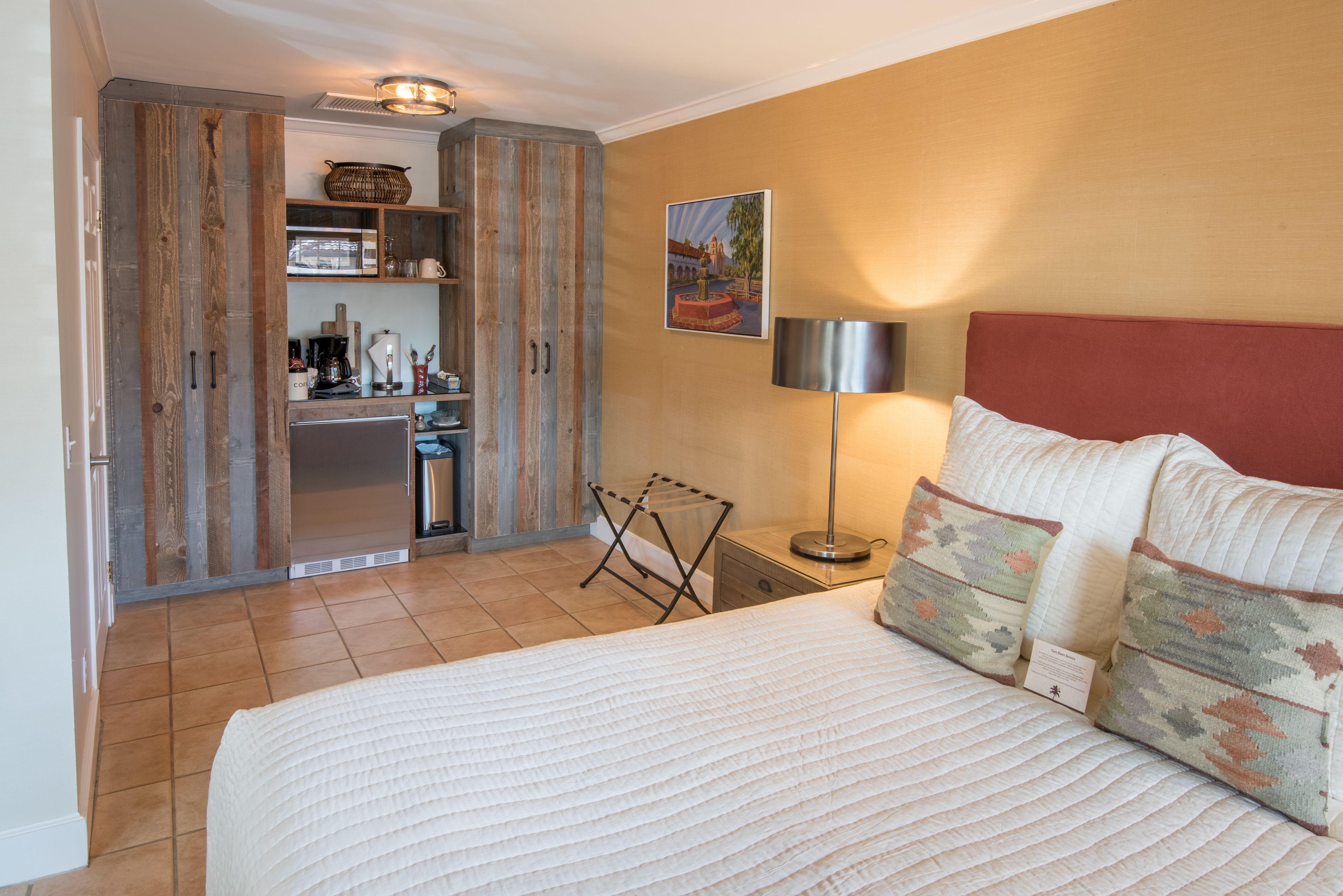 Quarto com cama, mesa de cabeceira, luminária, pintura na parede e cozinha compacta com armários e um cabideiro.