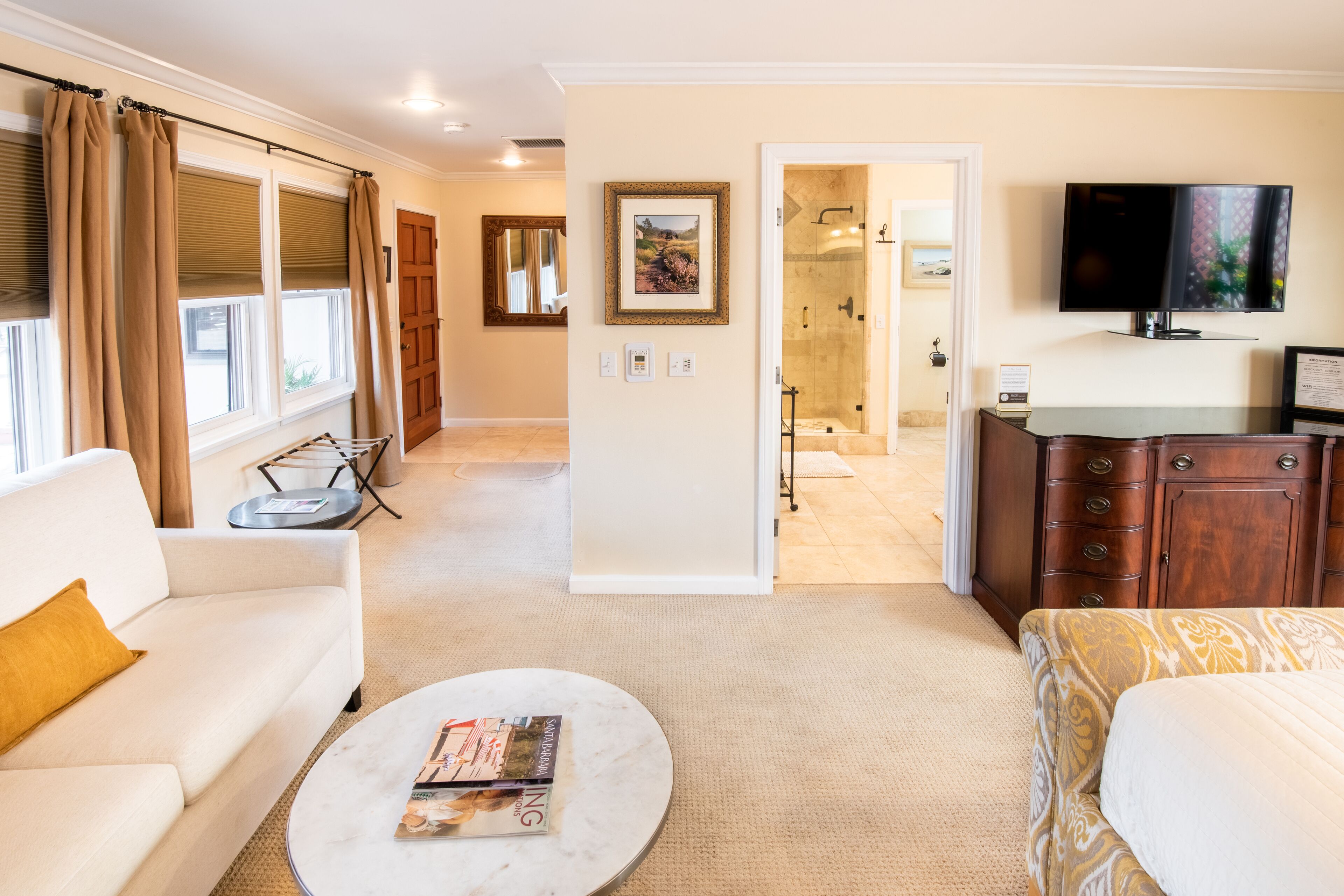 A hotel room with a sofa, a coffee table with magazines, a TV, a wardrobe, and a bathroom visible through an open door.