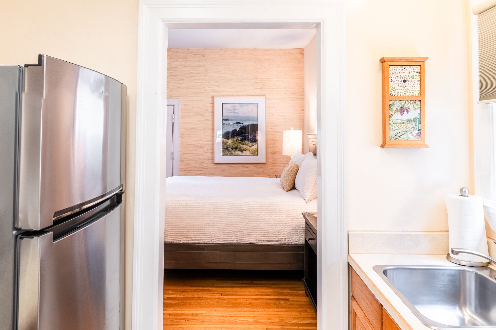 A compact hotel room with a bed, a small bedside table, a wall-mounted picture, a refrigerator, and a kitchen area with a sink and countertop.