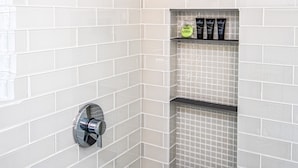 Une salle de bain avec des murs recouverts de carreaux blancs, une pomme de douche et une étagère contenant des articles de toilette.