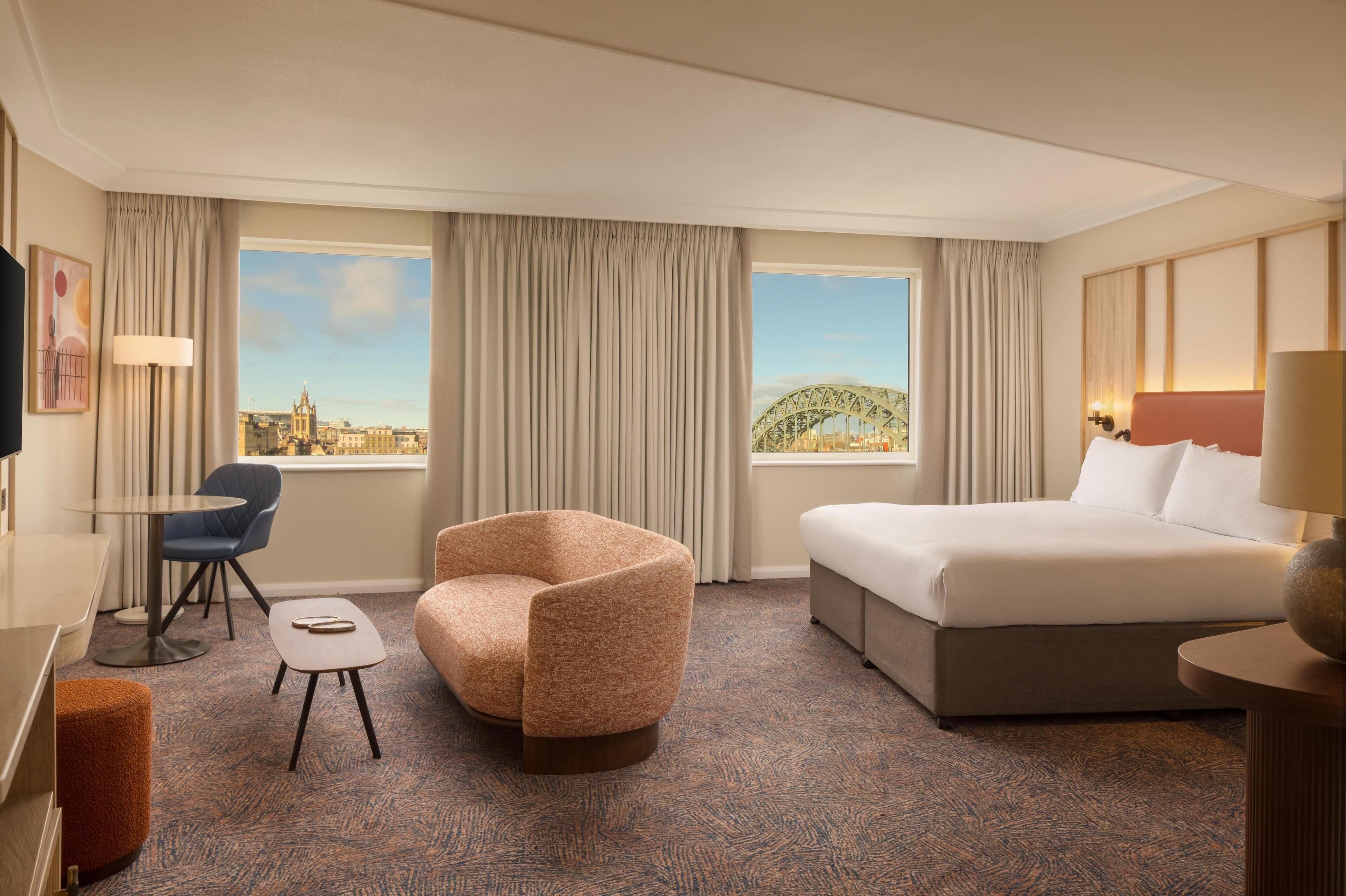 A hotel room with a bed, a chair, a small table, and a view of a cityscape and a bridge.
