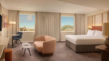 A hotel room with a bed, a chair, a small table, and a view of a cityscape and a bridge.