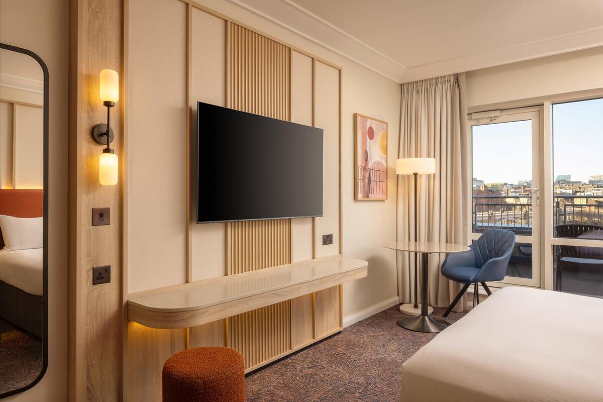 A hotel room with a large bed, a TV mounted on the wall, a small table, a chair, and a window with a city view.