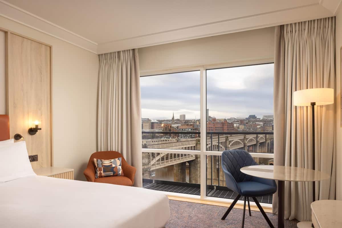 A hotel room with a bed, a chair, a small table, and a large window offering a city view.