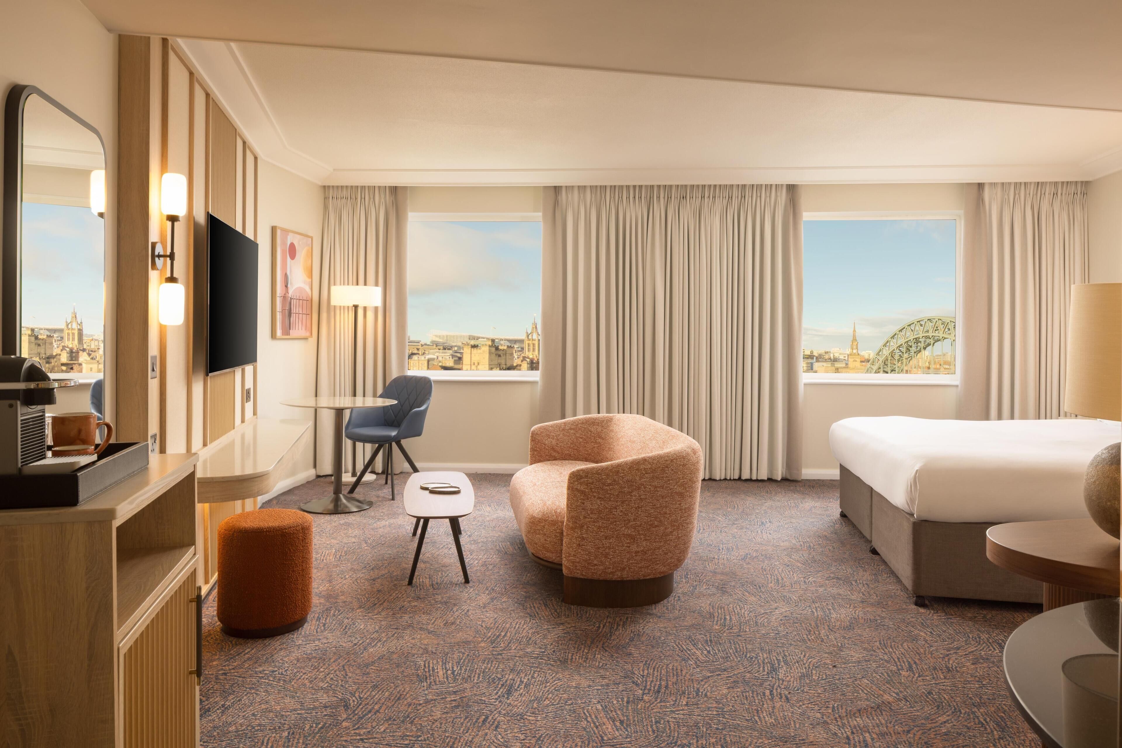 A hotel room with a bed, a sofa, a chair, a small table, a TV, and a view of a cityscape.