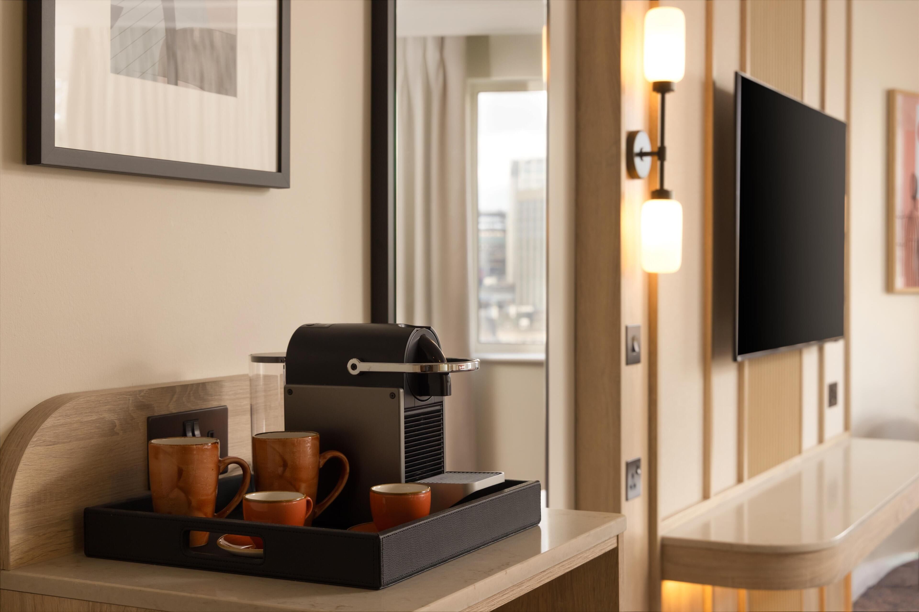 A hotel room with a coffee machine, mugs, a tray, a flat-screen TV, and a framed picture on the wall.