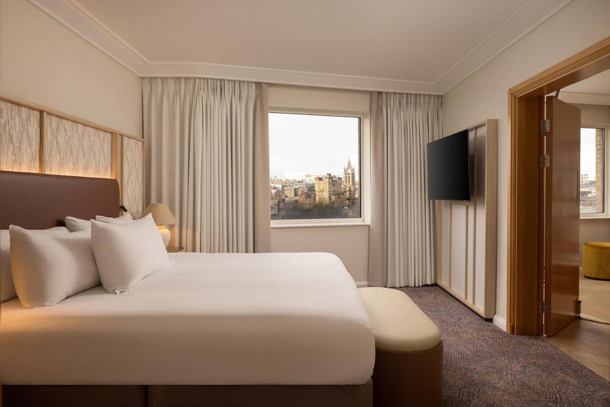 A hotel room with a large bed, a flat-screen TV, a window with curtains, and a view of the cityscape.