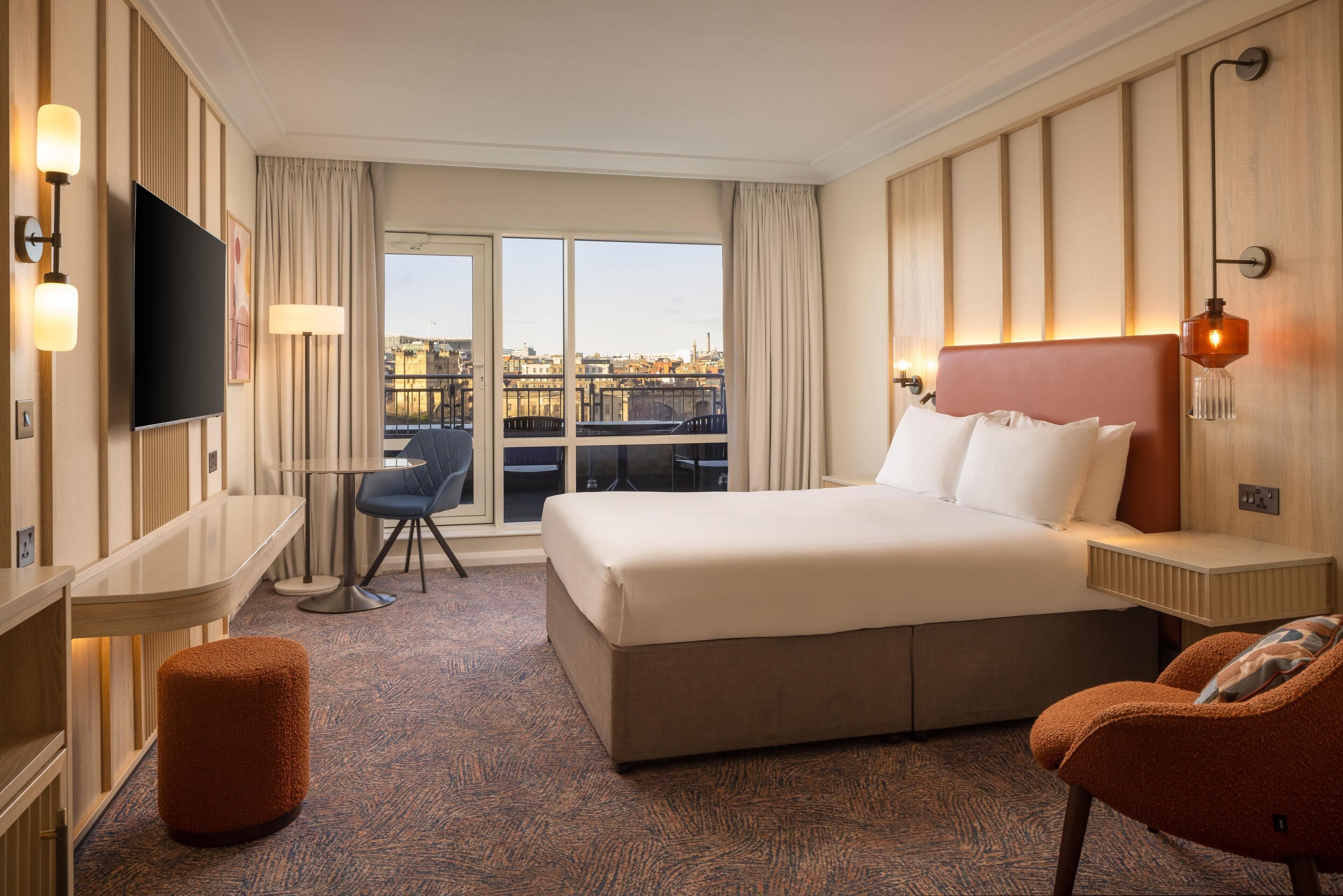 A hotel room with a large bed, a desk, a chair, a television, and a view of the city through the windows.