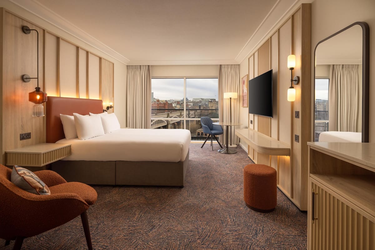 A hotel room with a large bed, a desk, a chair, a TV, and a view of the city.