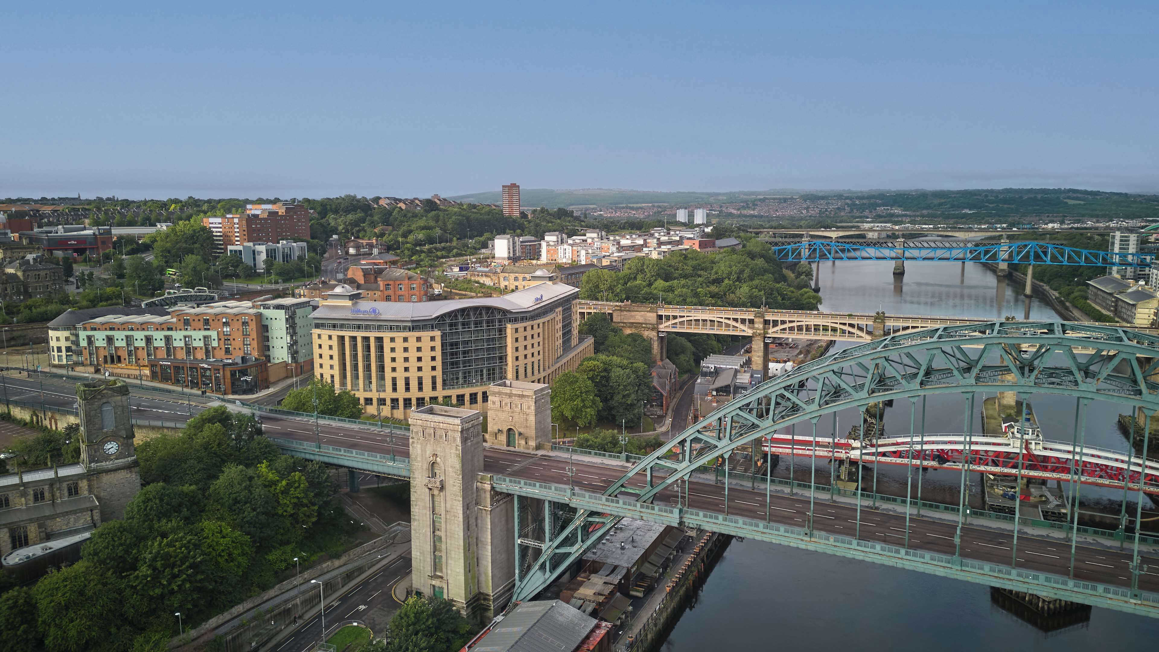 Un paisaje urbano con un puente verde destacada que cruza un río, rodeado de diversos edificios, incluyendo una gran estructura amarilla y una torre de reloj histórica.