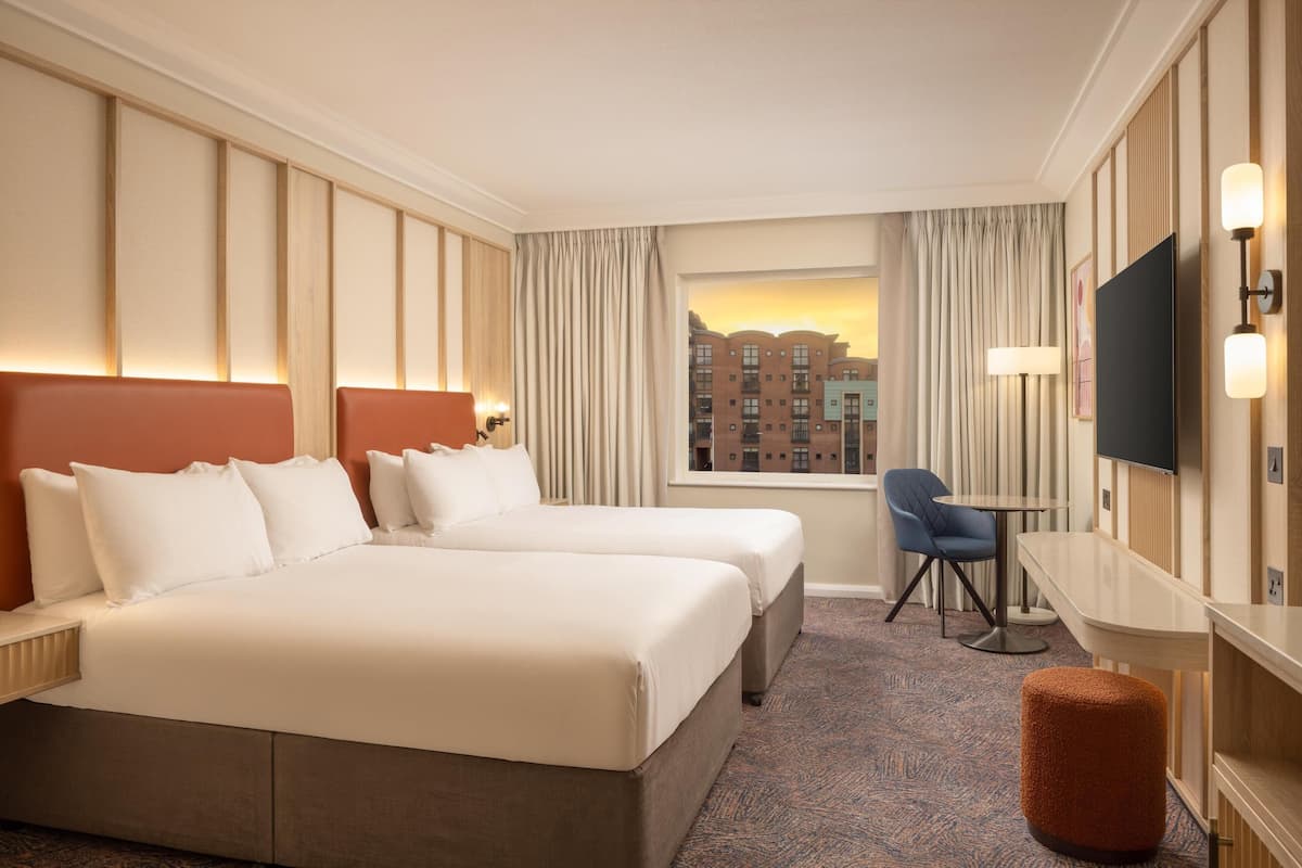 A hotel room with two beds, a desk, a chair, a TV, and a window with curtains.