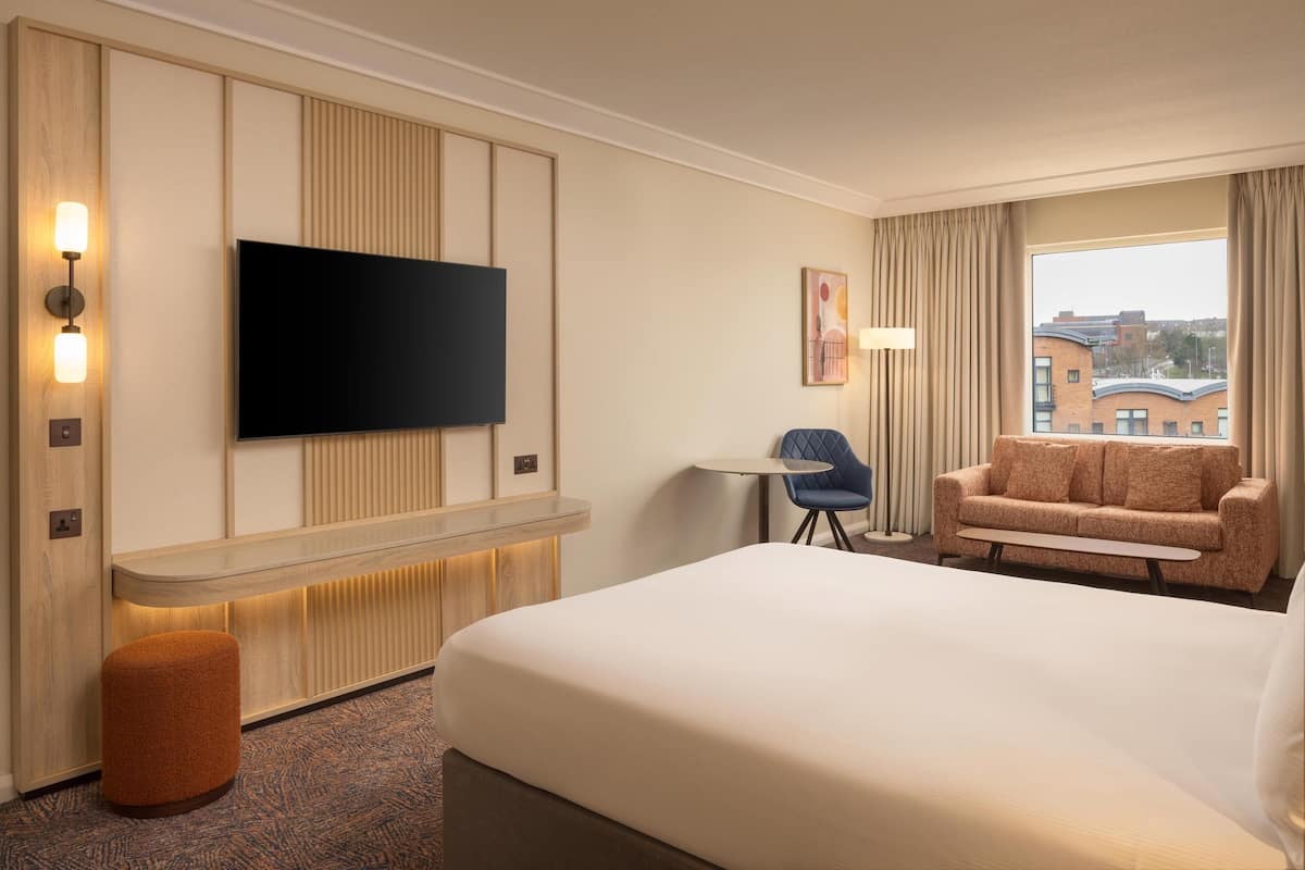 A hotel room with a large bed, a flat-screen TV mounted on the wall, a small table, a blue chair, and a sofa.
