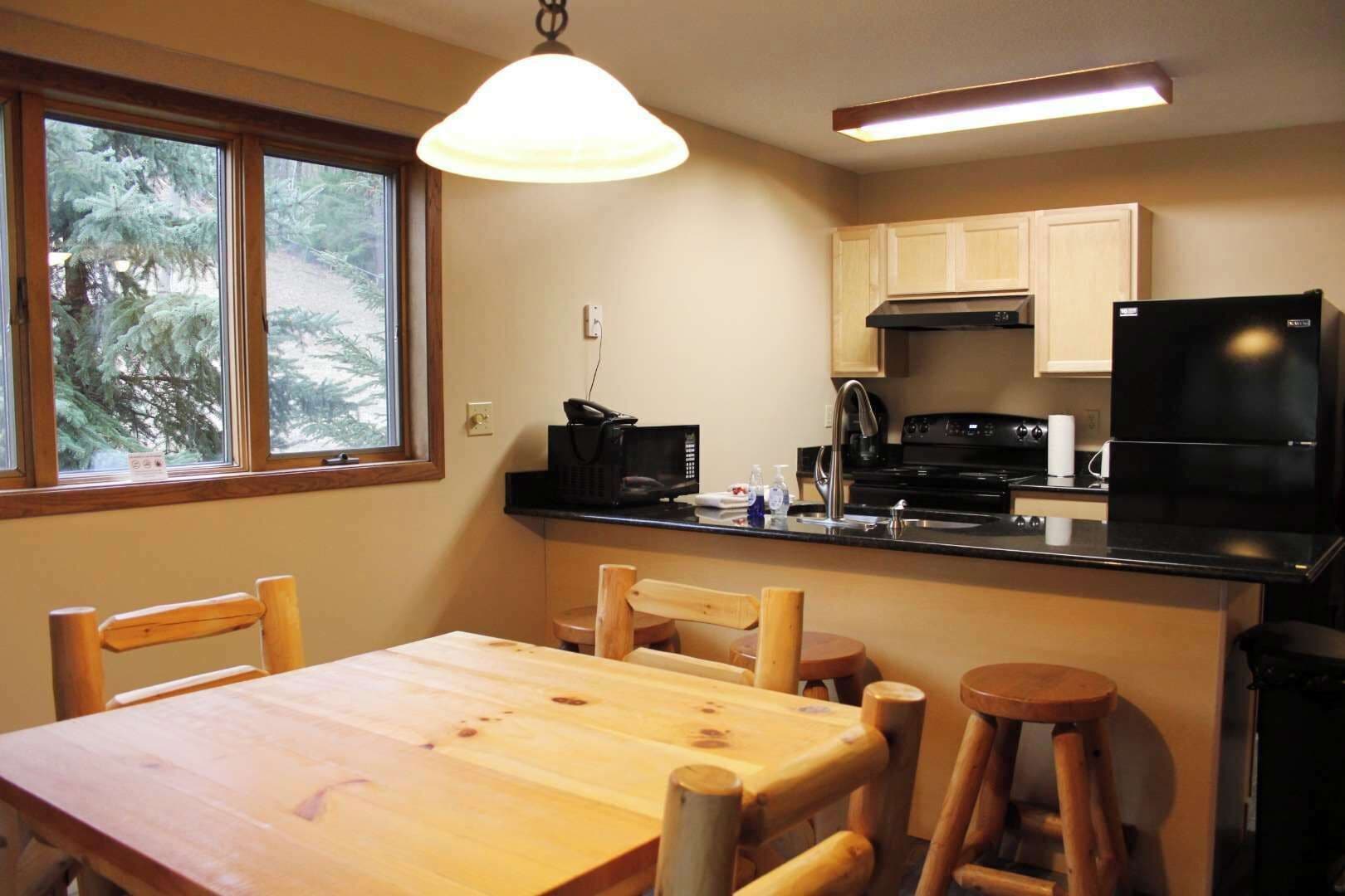 Una cocina compacta con una mesa de madera y taburetes, un refrigerador negro y una ventana con vista a árboles.