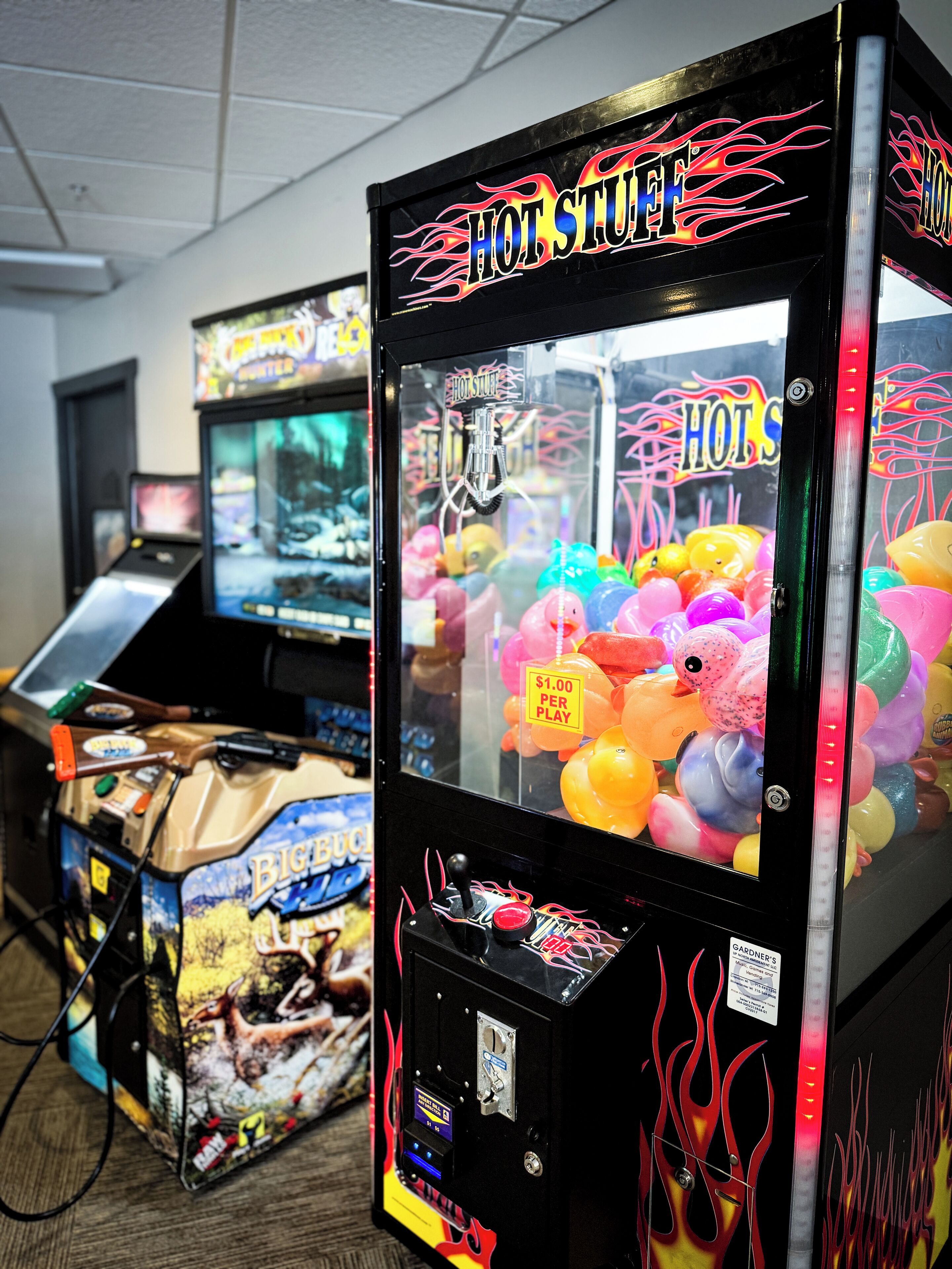 Une rangée de jeux d’arcade, dont un jeu de griffe intitulé « Hot Stuff » et un jeu de tir intitulé « Big Buck Hunter », dans une pièce dont le sol est recouvert d’une moquette et dont le plafond est blanc.