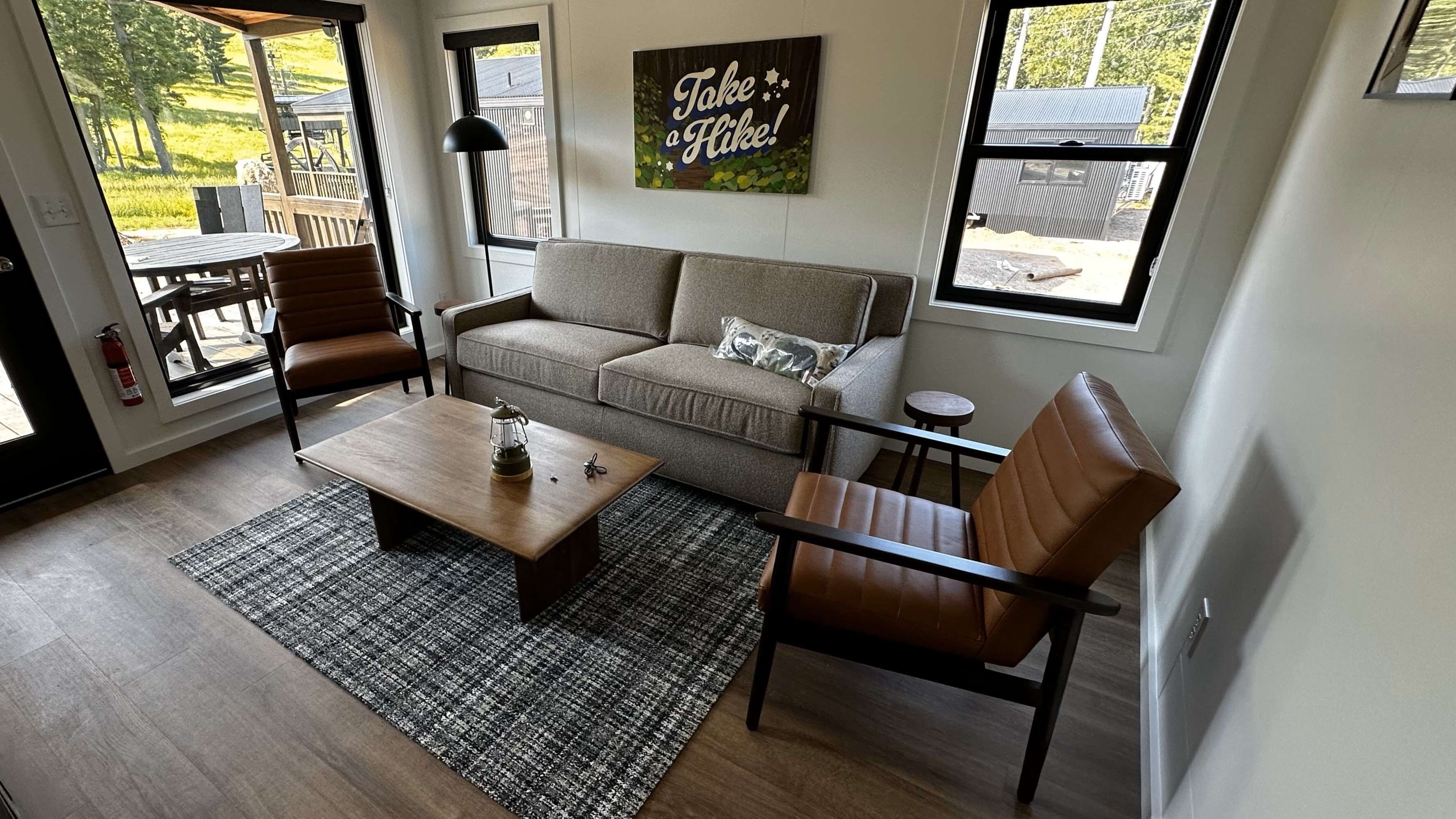 A modern living room with a sofa, two chairs, a coffee table, and a rug. There is a painting on the wall and a window with a view of the outdoors.