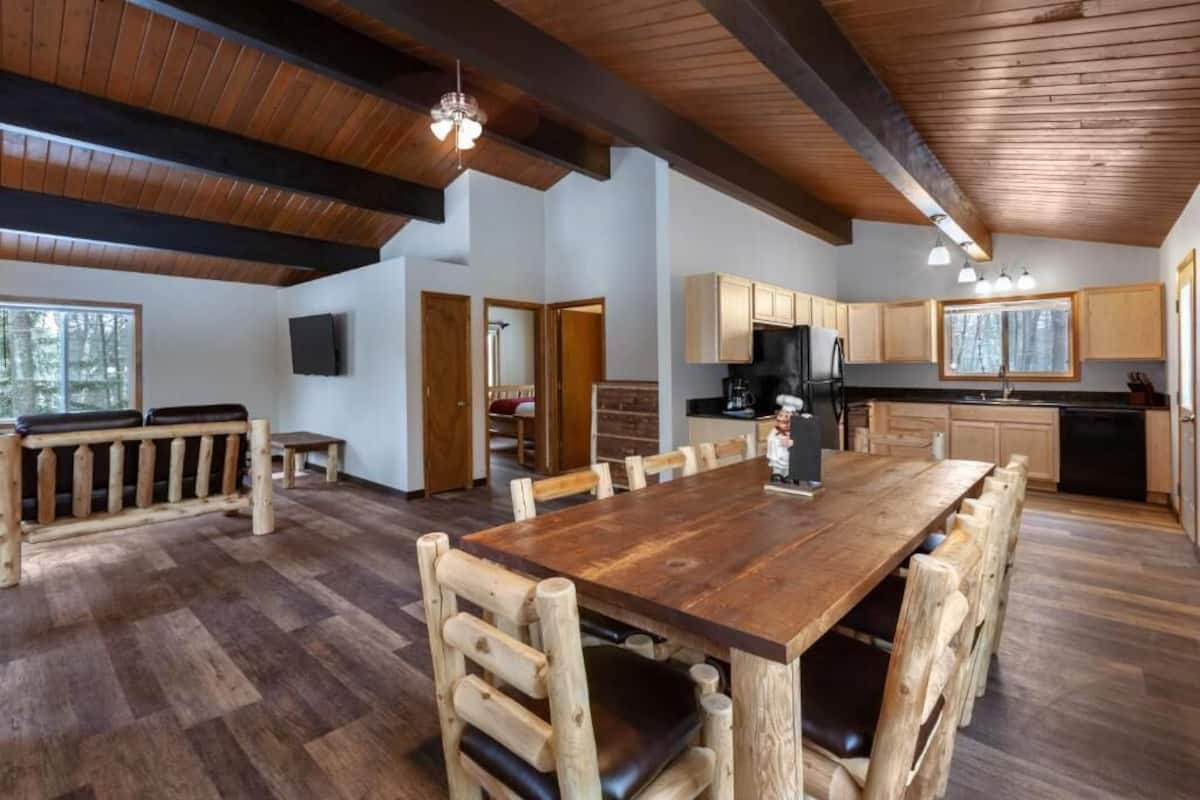 A spacious dining area with a large wooden table and chairs, a kitchen with wooden cabinets, and a view of the forest through large windows.