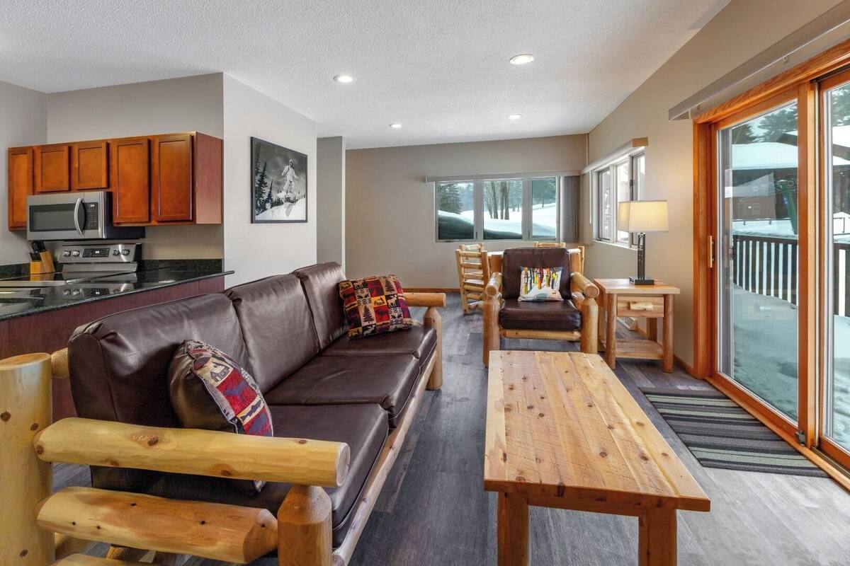 A cozy living room with a leather sofa, wooden armchairs, a wooden coffee table, and a kitchen area with a microwave and stove.