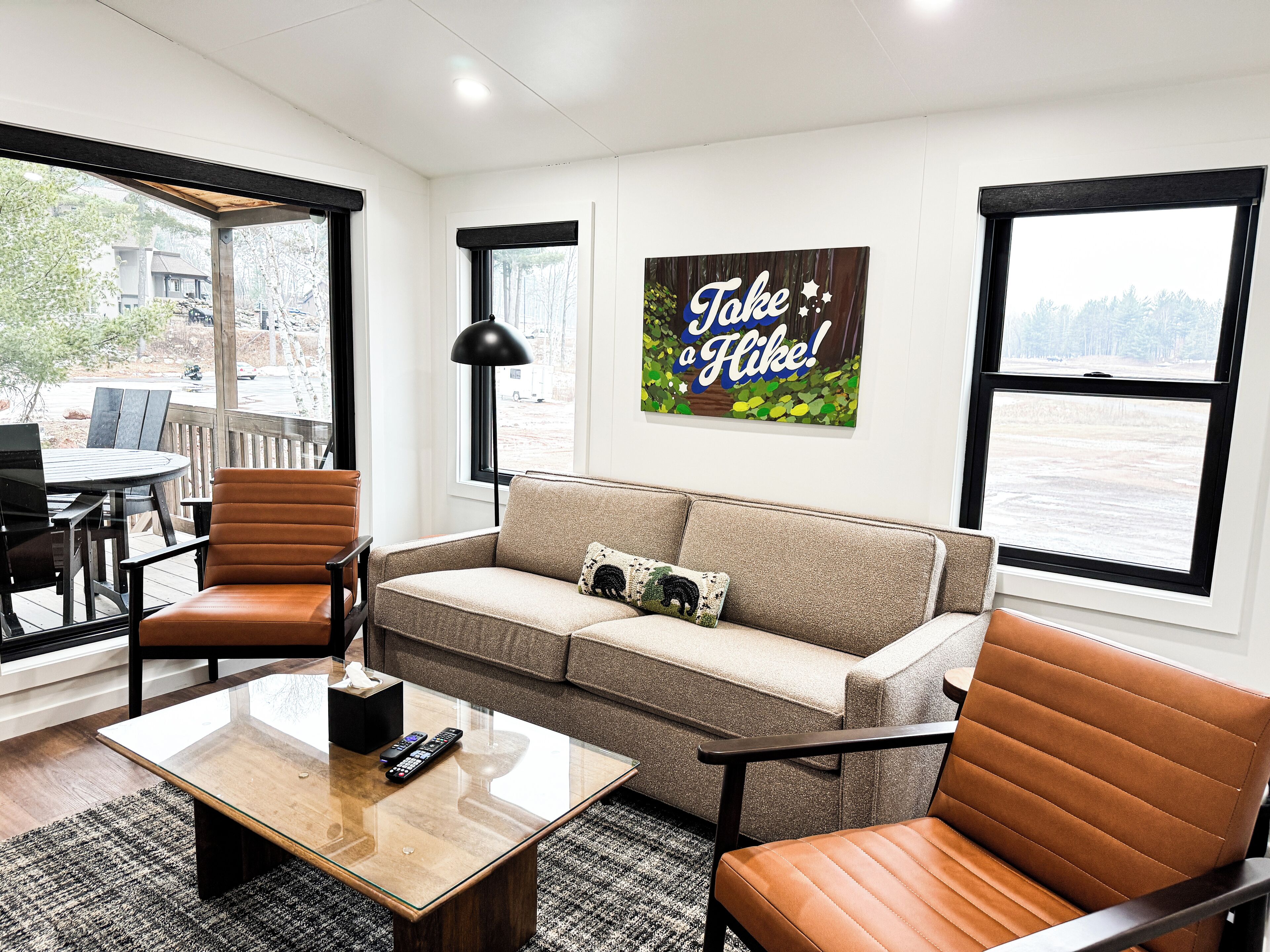 A living room with a sofa, two armchairs, a coffee table, and a TV. There are two windows with a view of trees and a sign that says "Take a Hike!"