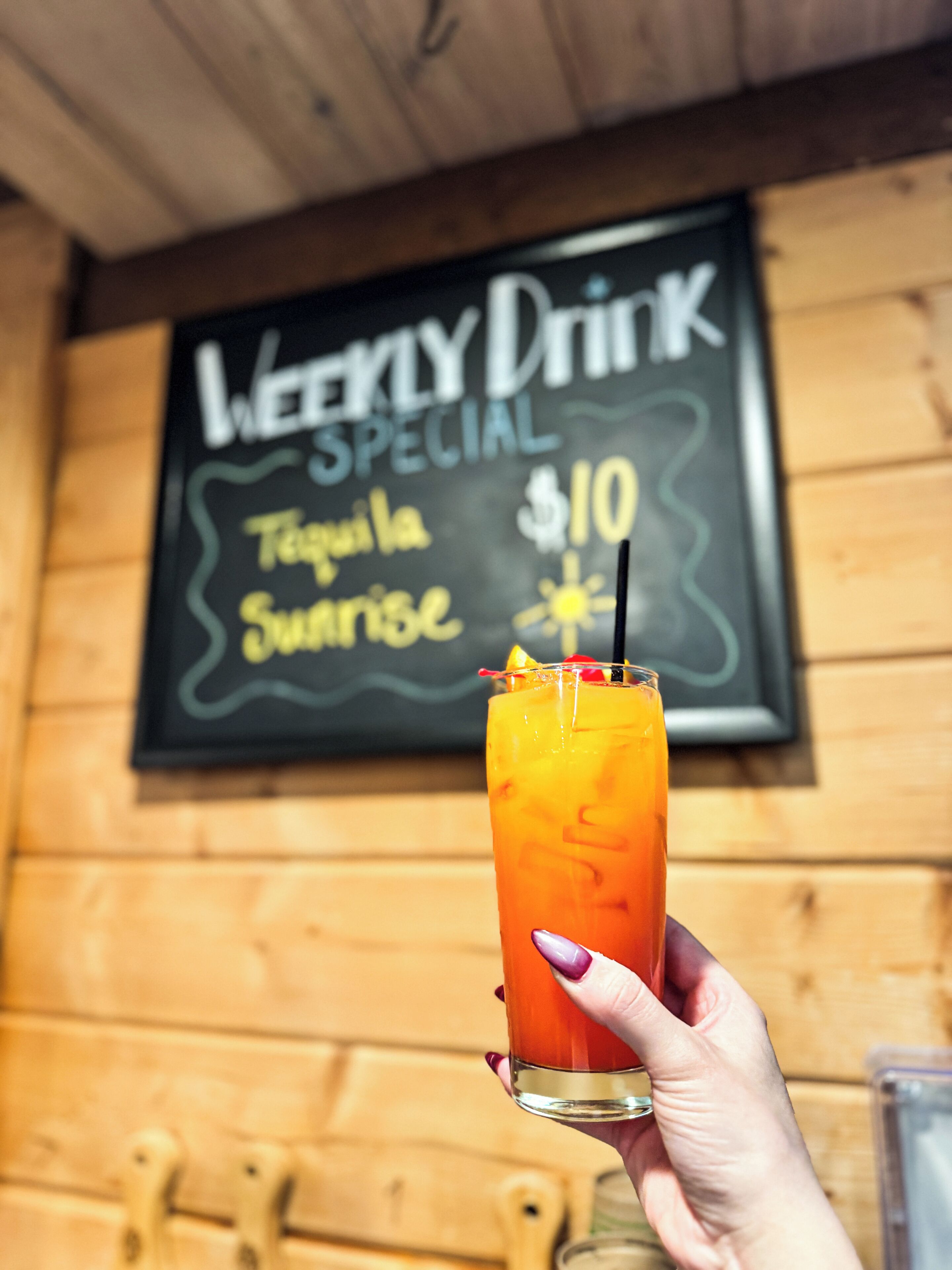 A hand holding a glass of a yellow beverage with a straw, in front of a chalkboard sign advertising a special drink for $10.