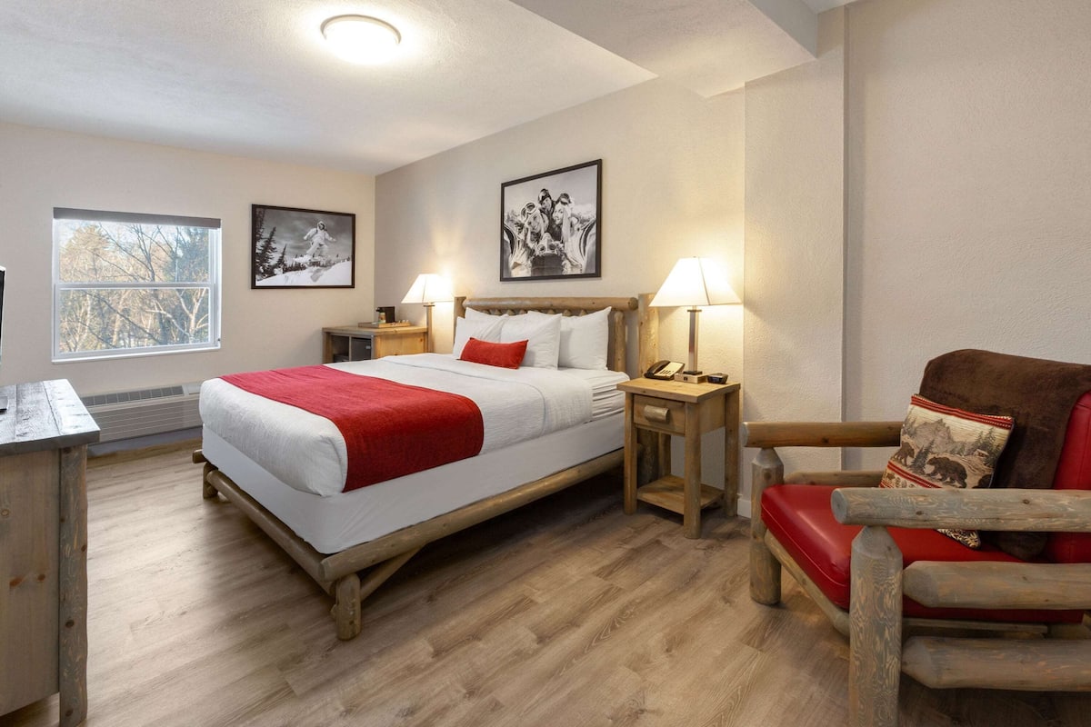 A bedroom with a wooden bed, a red chair, a nightstand with a lamp, and a window with a view of trees.