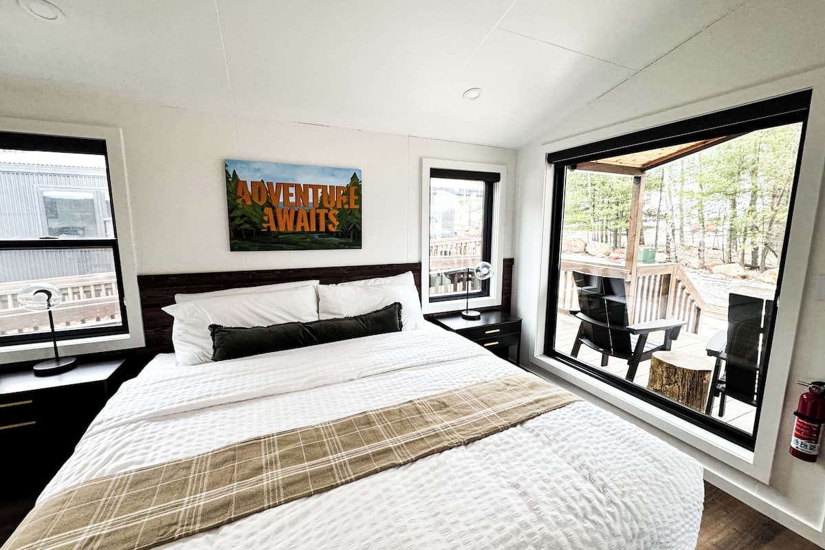 A modern hotel room with a large bed, a painting, and a view of a deck and trees.