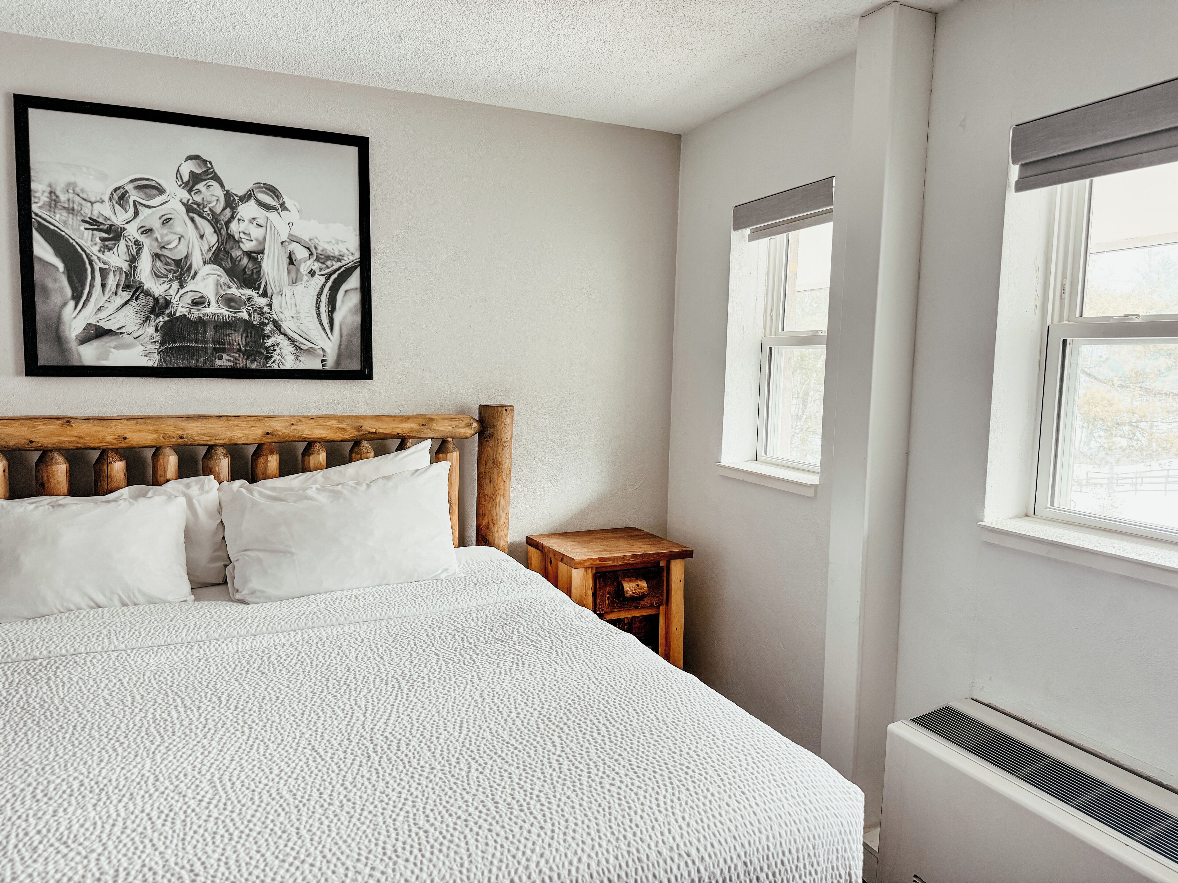 A bedroom with a wooden headboard, white bedding, a small wooden nightstand, and a framed photograph on the wall.