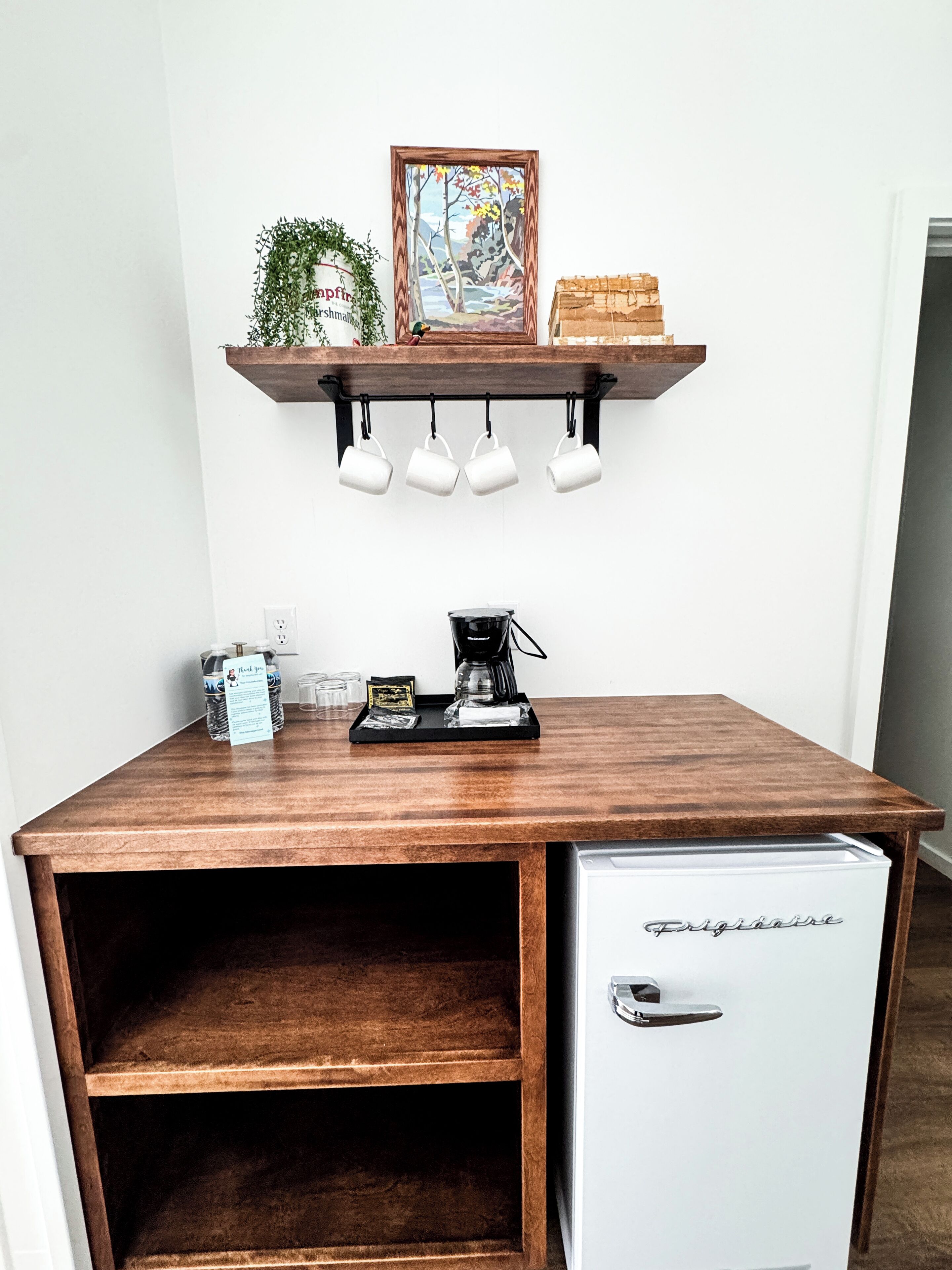 A compact kitchenette with a coffee maker, a small fridge, and a shelf above holding mugs and a framed picture.