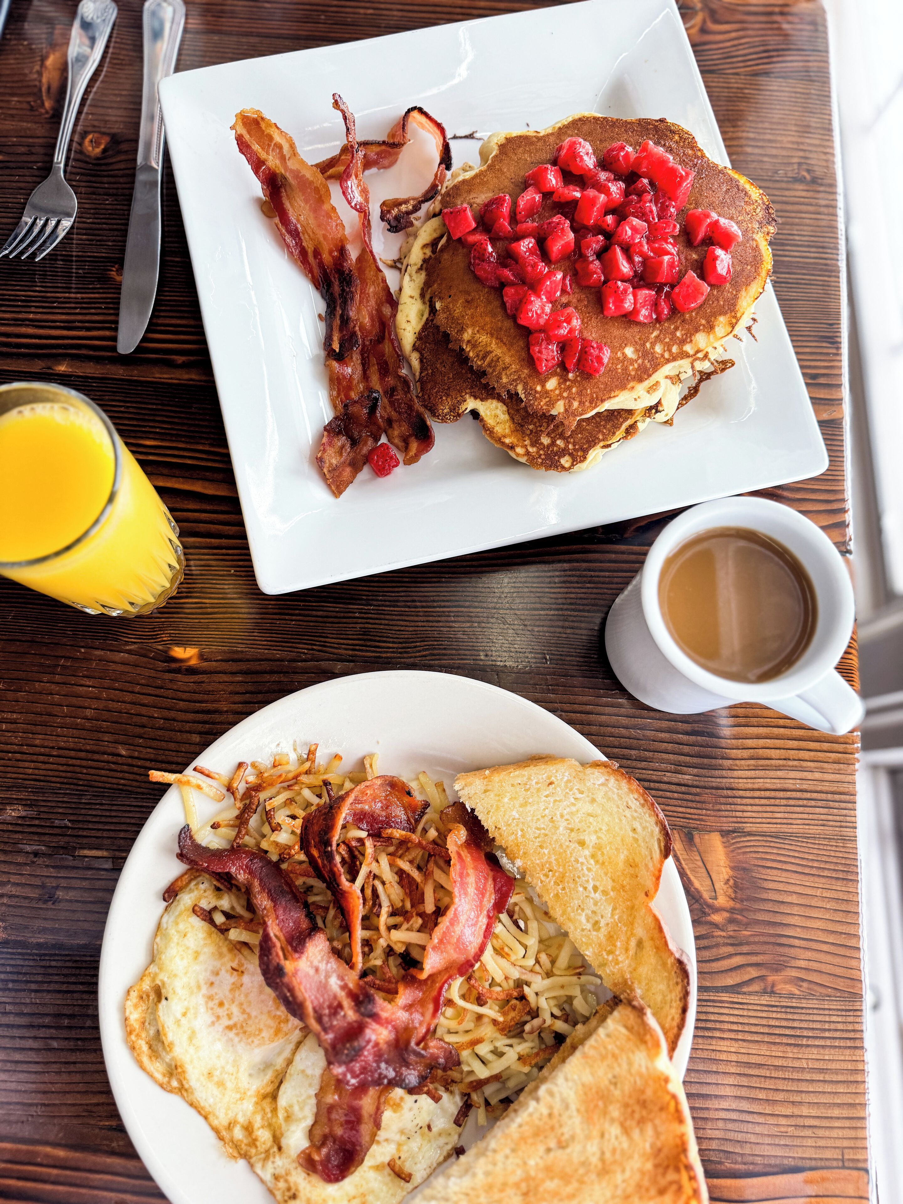 A breakfast plate with pancakes topped with red berries, bacon, and hash browns, alongside a glass of orange juice and a cup of coffee on a wooden table.