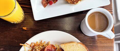 A breakfast plate with pancakes topped with red berries, bacon, and hash browns, alongside a glass of orange juice and a cup of coffee on a wooden table.