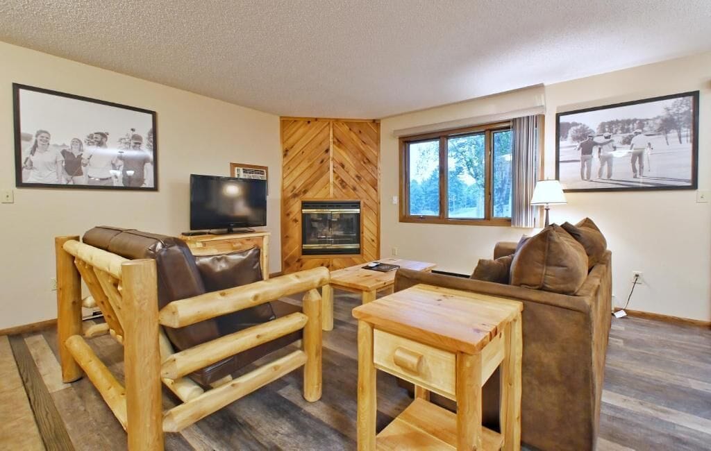 A cozy living room with a fireplace, wooden furniture, and large windows.
