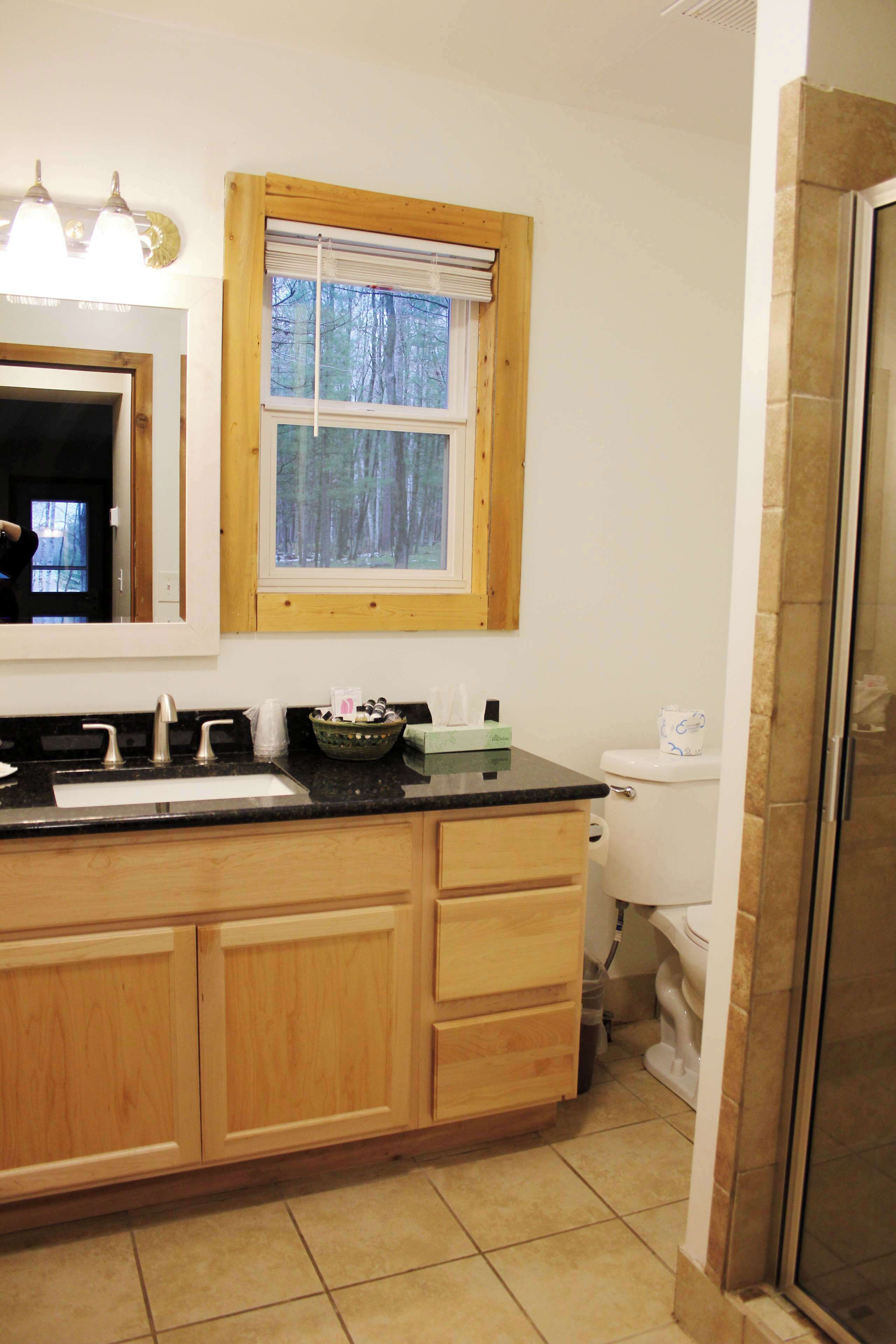 A bathroom with a wooden vanity, a sink, a mirror, a window with blinds, and a toilet.