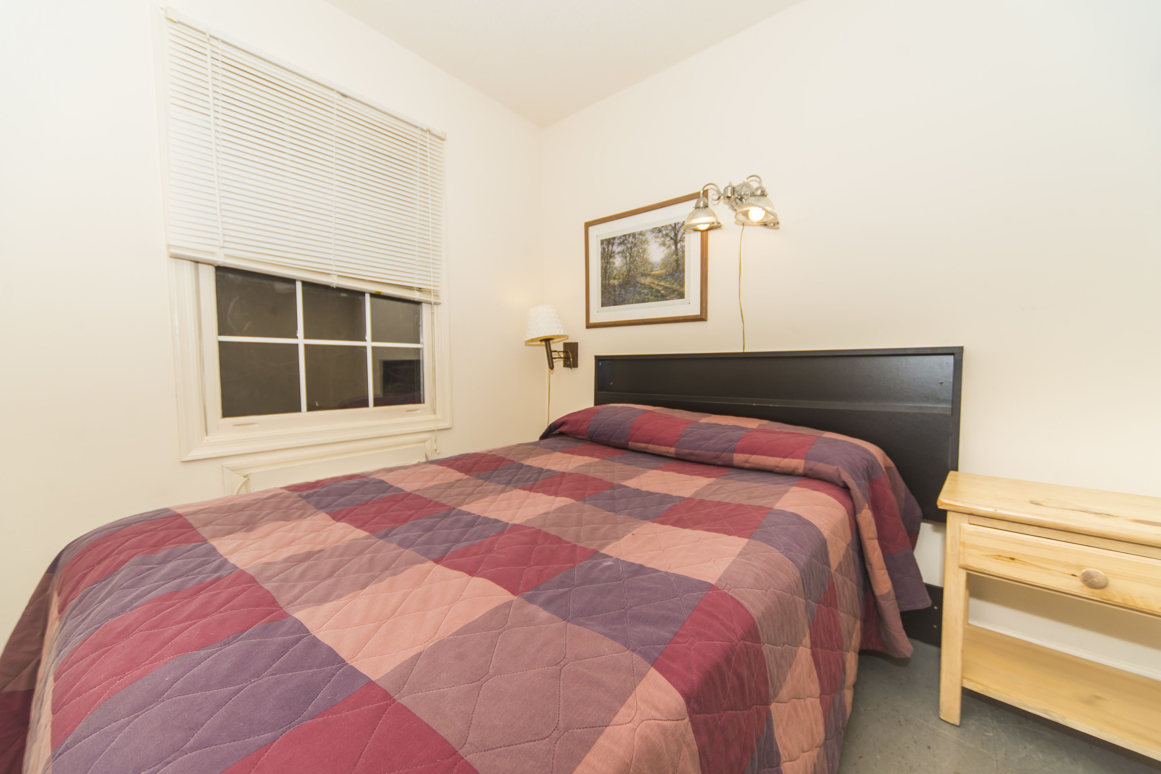 Ein Schlafzimmer mit Bett, Nachttisch, Fenster mit Jalousien und einer Wandlampe.