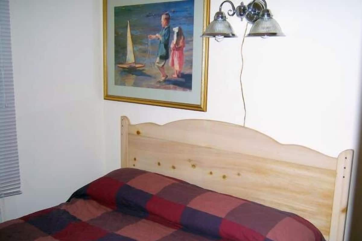 A bed with a wooden headboard, a checkered bedspread, and a painting of a sailboat on the wall.
