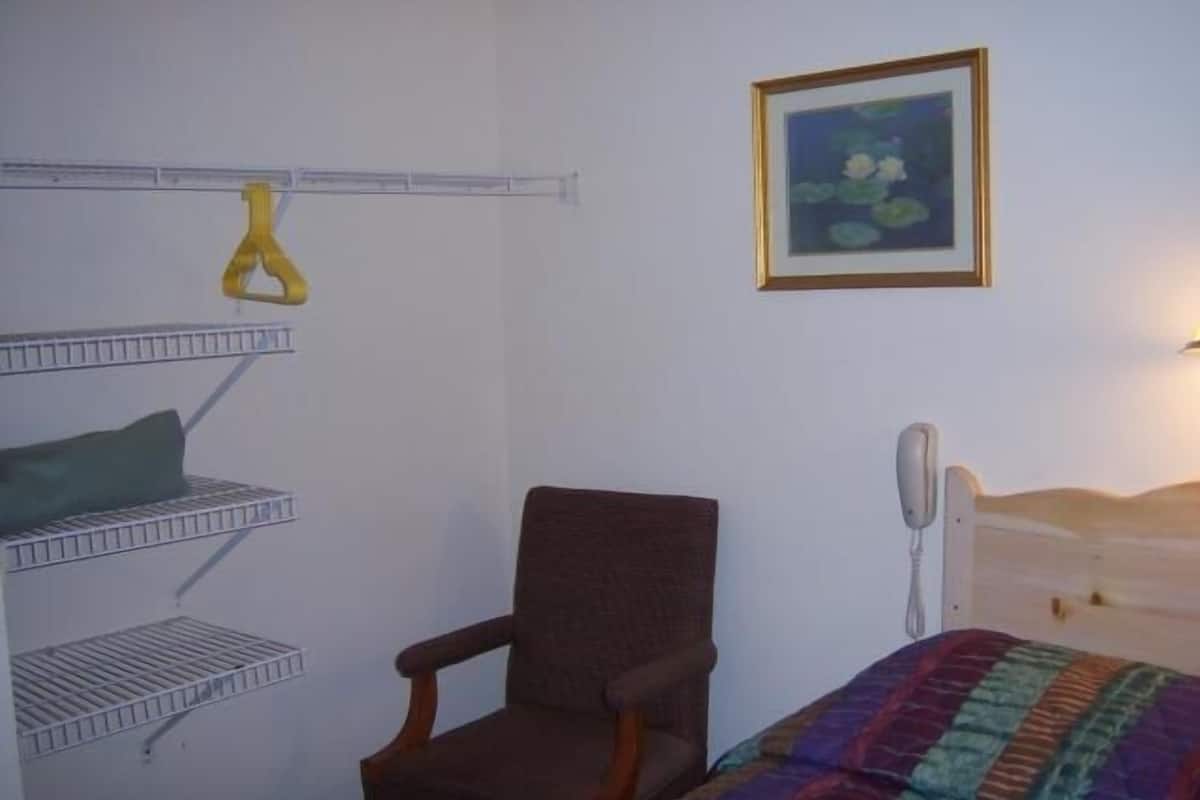 A bedroom with a bed, a chair, a shelf with a yellow item, a framed picture, and a bedside telephone.